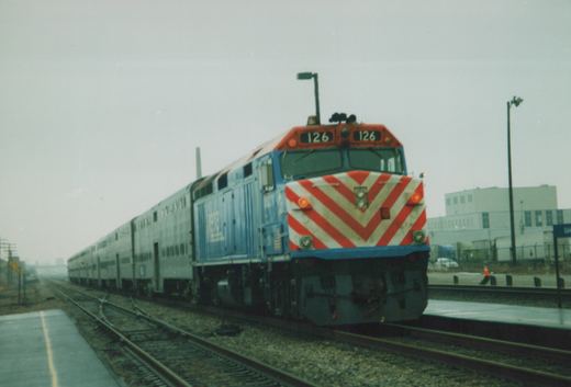 A blue Metra diesel locomotive, front number 126, is shown at a station platform with its train of silver commuter cars behind it on a cloudy day.