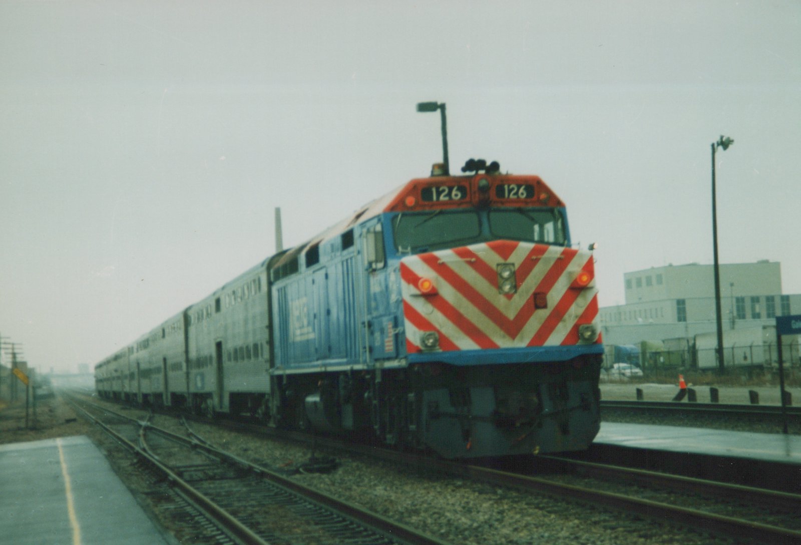 A blue Metra diesel locomotive, front number 126, is shown at a station platform with its train of silver commuter cars behind it on a cloudy day.