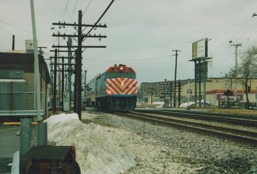 A blue, gray, and red locomotive with a chevron front and the number 126 on its front travels on railroad tracks in a suburban industrial area with snow piles and utility poles.