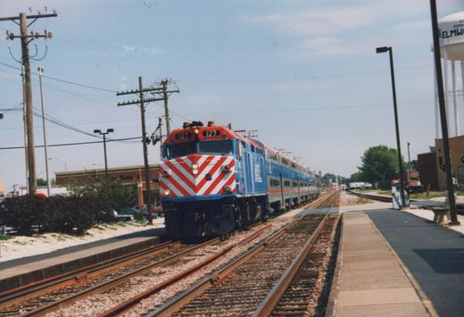 A blue and red Metra train with locomotive number 127 and gallery cars is stopped at a station platform with a water tower reading 'ELMWO' in the background.