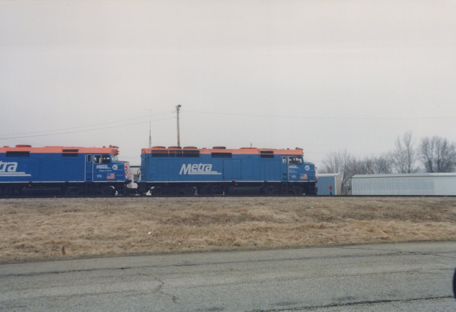 Two blue and orange Metra diesel locomotives, numbered 131 and 175, are parked next to each other on a railway track with bare trees in the background.