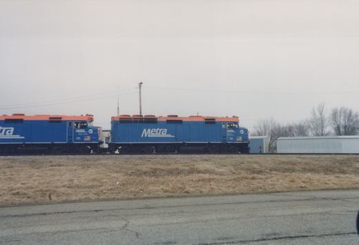 Two blue and orange Metra diesel locomotives, numbered 131 and 175, are parked next to each other on a railway track with bare trees in the background.