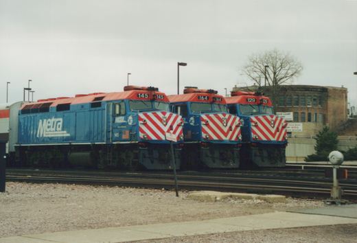 Three blue, red, and white Metra diesel locomotives with numbers 145, 164, and 129 are parked together on railroad tracks in a yard with a building and bare trees in the background.