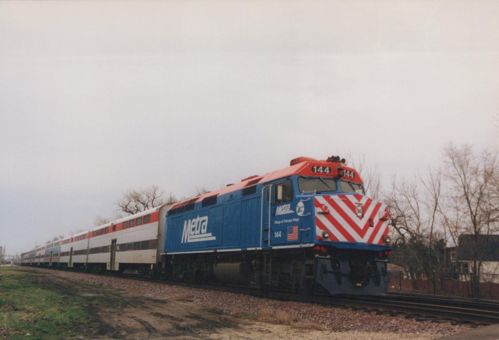 A blue and red Metra locomotive numbered 144 pulls a long train of white commuter cars on a cloudy winter day, with bare trees and a fence in the background.