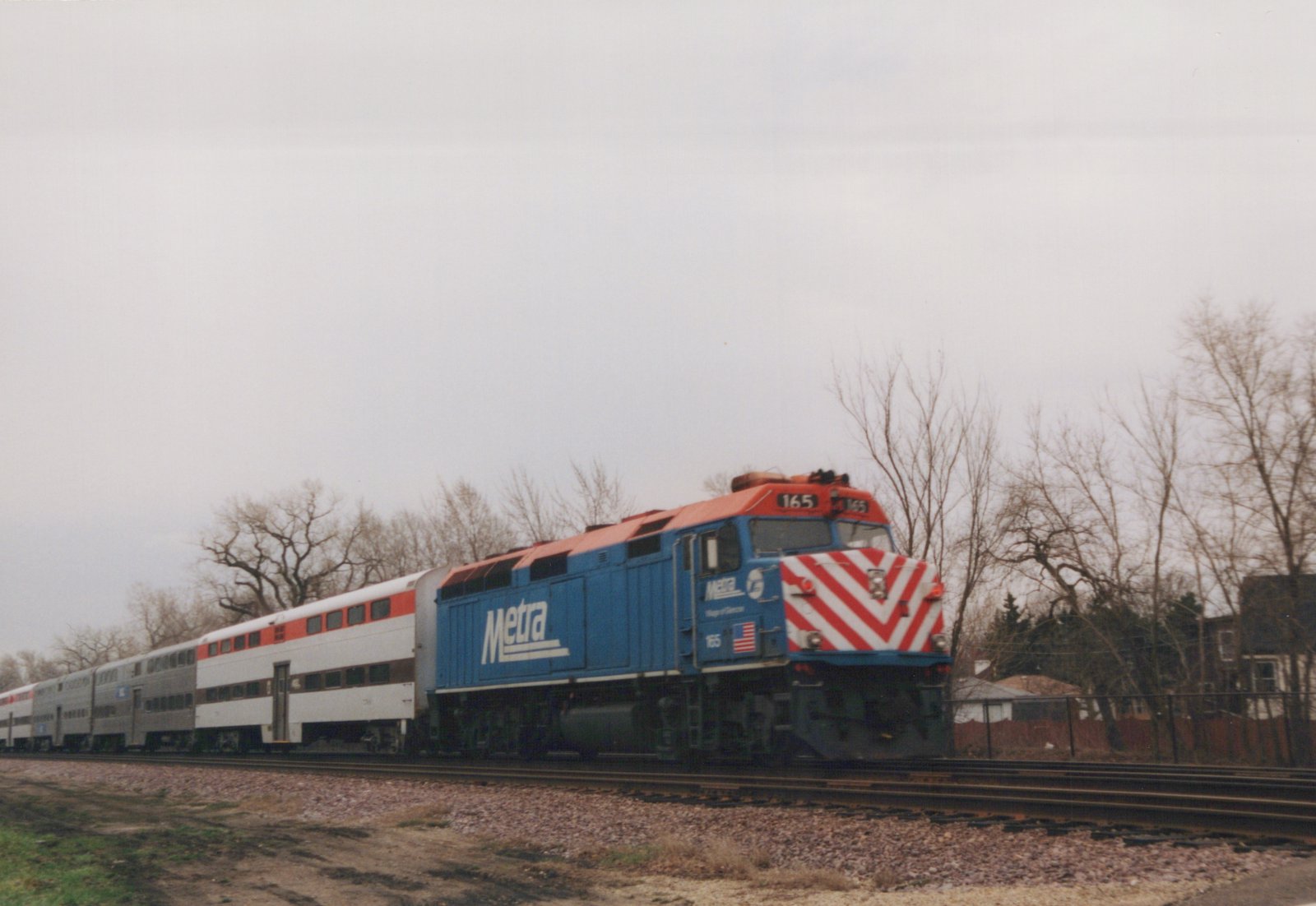 A blue and red Metra locomotive with the number 165 leads a train of double-decker commuter cars along a track lined with bare trees under an overcast sky.