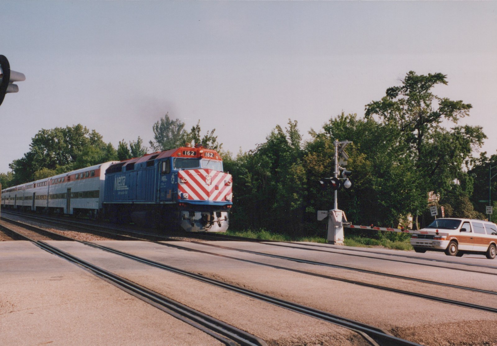 A blue Metra locomotive with red and white chevrons on its nose, numbered 162, pulls silver bi-level passenger cars across a road at a grade crossing on a sunny day, with trees in the background.