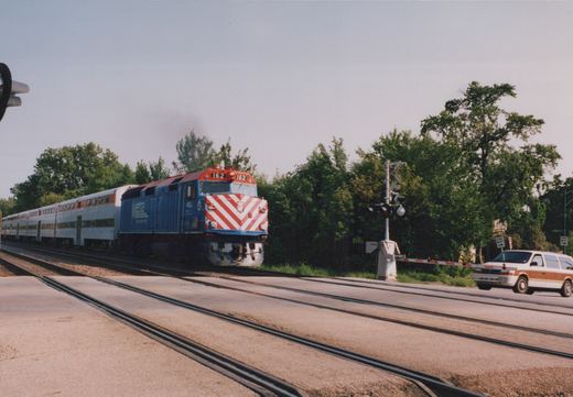 A blue Metra locomotive with red and white chevrons on its nose, numbered 162, pulls silver bi-level passenger cars across a road at a grade crossing on a sunny day, with trees in the background.