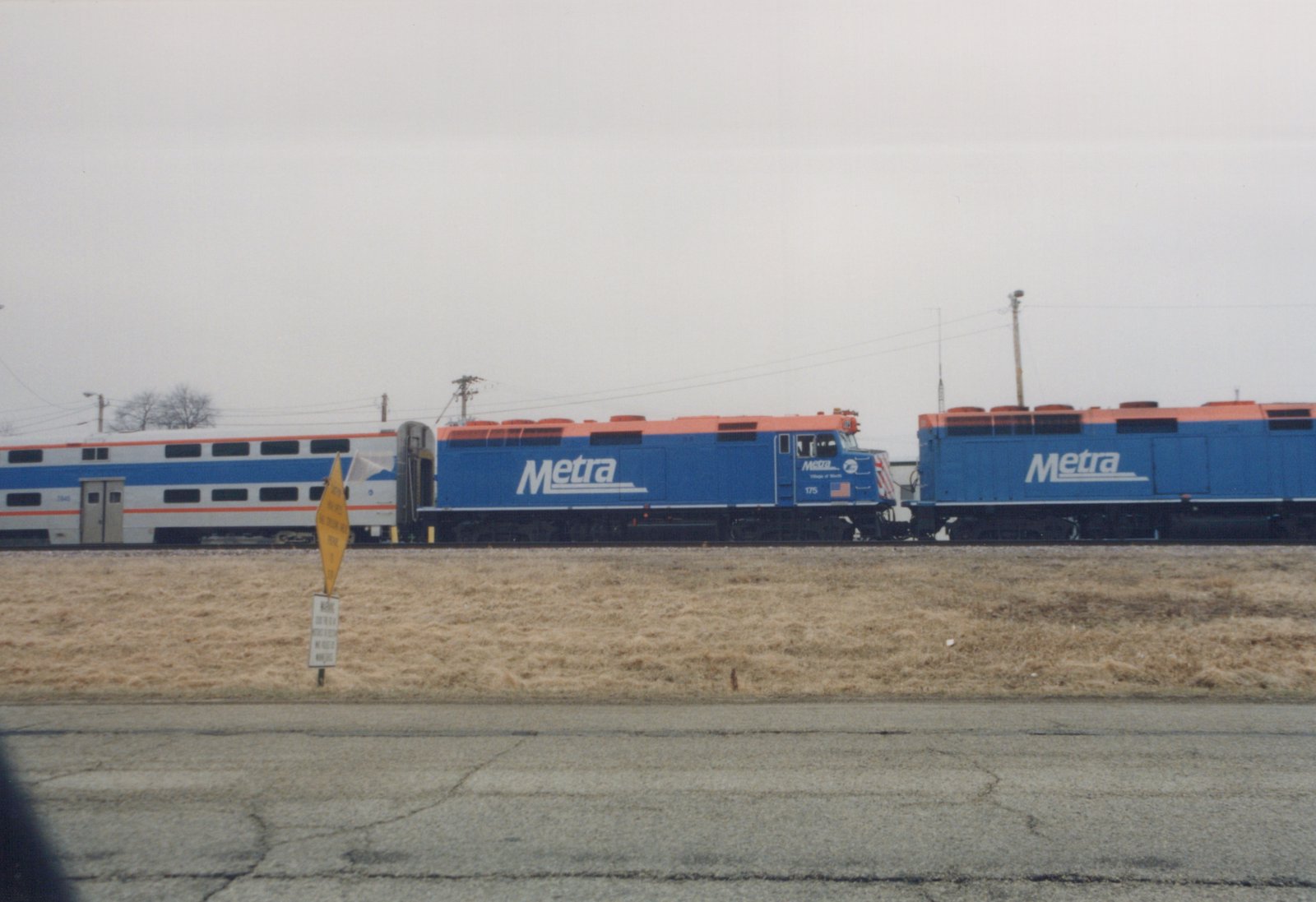 A side view of a blue and orange Metra train with locomotive 175 pulling a passenger car, with a second locomotive behind, on a track next to a road with dry grass in the foreground under an overcast sky.