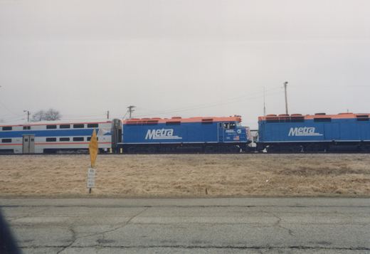 A side view of a blue and orange Metra train with locomotive 175 pulling a passenger car, with a second locomotive behind, on a track next to a road with dry grass in the foreground under an overcast sky.
