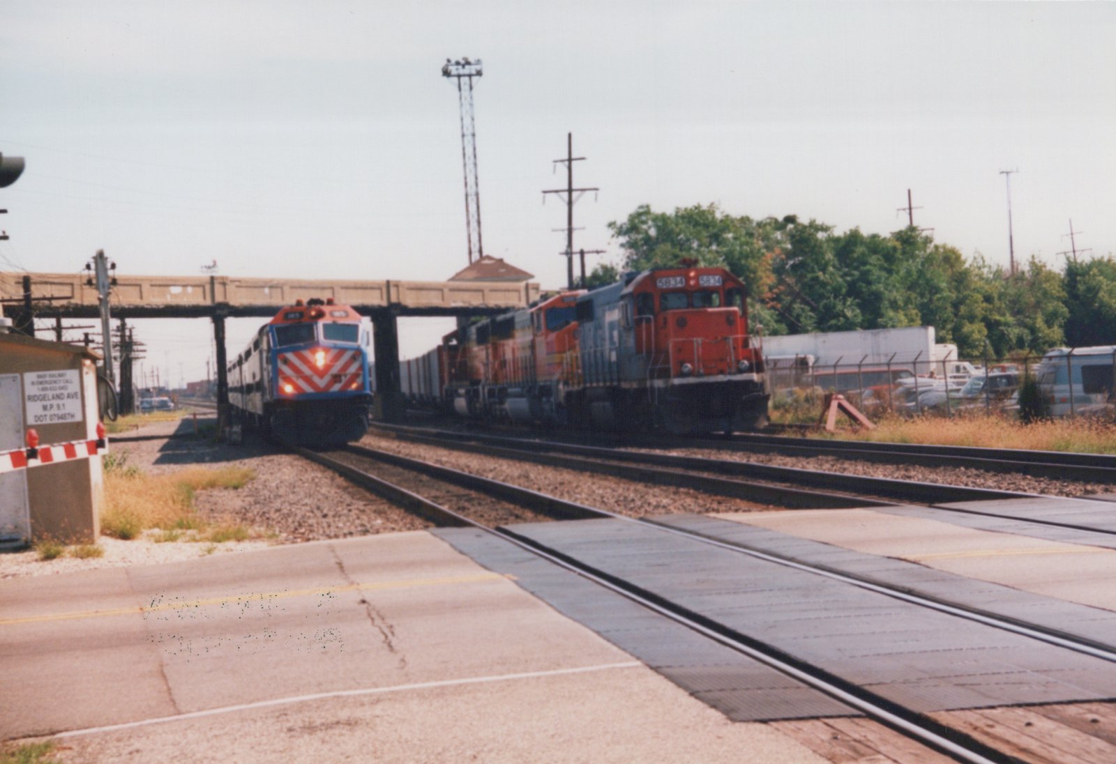 Two locomotives, one from BNSF Railway and one from Metra, travel on adjacent tracks near a grade crossing in a suburban setting.