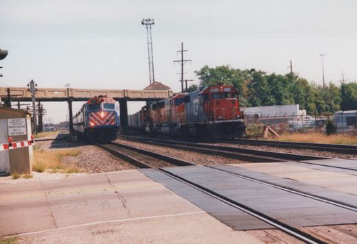 Two locomotives, one from BNSF Railway and one from Metra, travel on adjacent tracks near a grade crossing in a suburban setting.