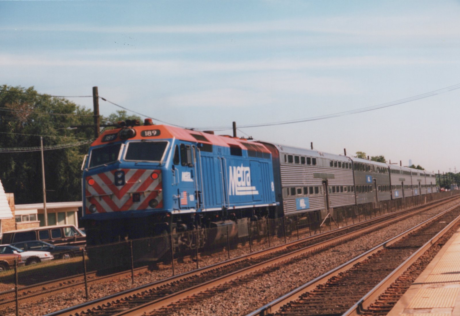 A blue and orange Metra locomotive numbered 189 pulls a long train of silver passenger cars on a sunny day, viewed from a station platform.