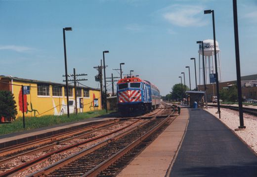A blue, silver, and red Metra locomotive numbered 212 travels along tracks past a yellow building and a water tower labeled 'Elmwood' at a station platform.