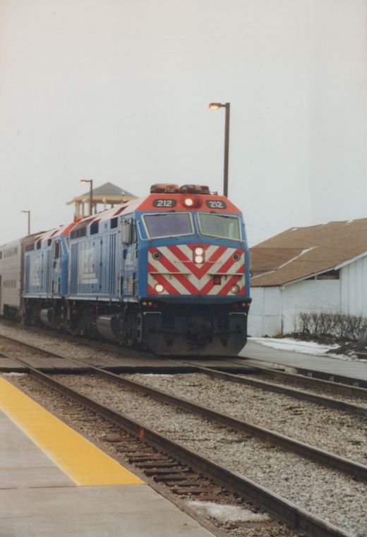 A blue and red Metra locomotive, number 212, is shown moving on the tracks next to a station platform with snow on the ground.
