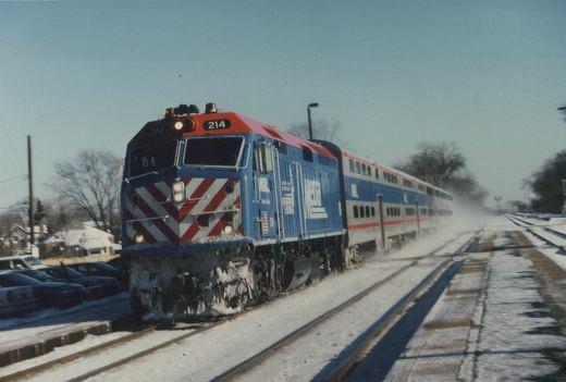 A blue and red Metra diesel locomotive pulling double-decker commuter cars on snow-covered tracks.