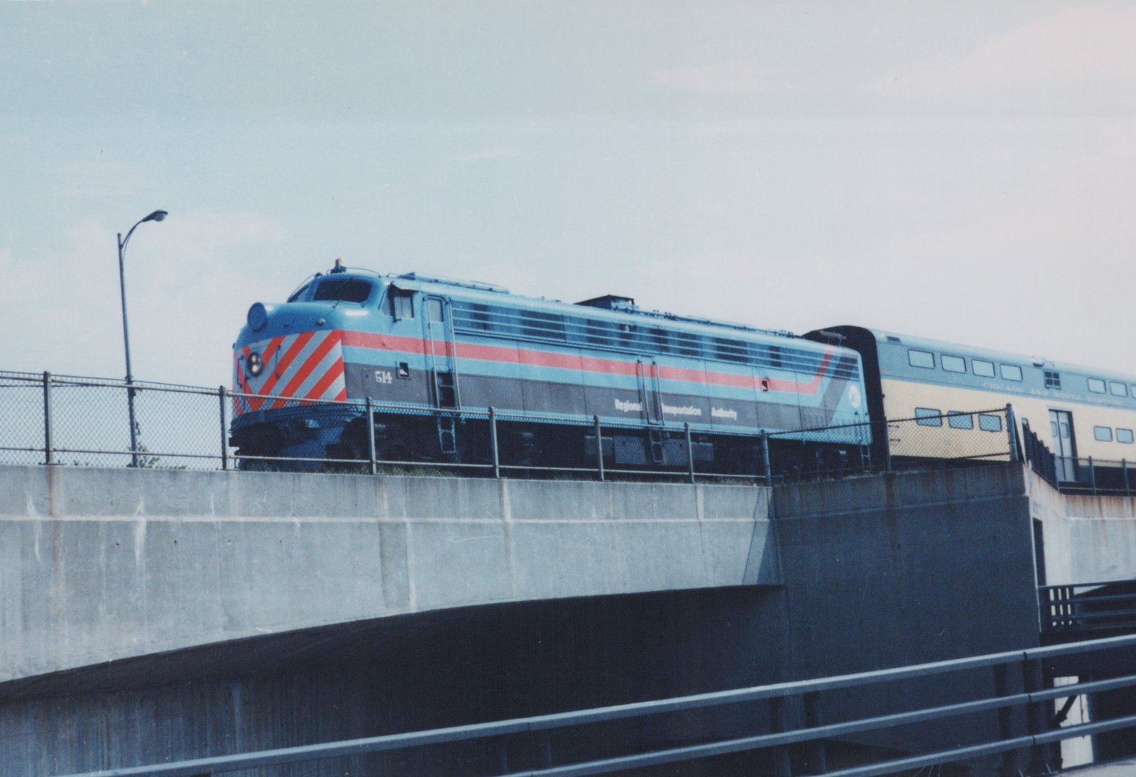 A blue, red and gray Regional Transportation Authority F40PH locomotive numbered 514 pulls a yellow and blue Chicago and North Western passenger car over a concrete viaduct under an overcast sky.