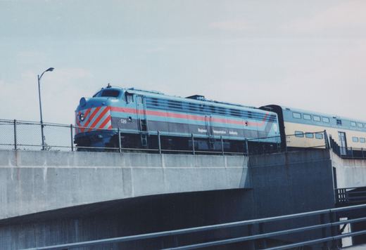 A blue, red and gray Regional Transportation Authority F40PH locomotive numbered 514 pulls a yellow and blue Chicago and North Western passenger car over a concrete viaduct under an overcast sky.