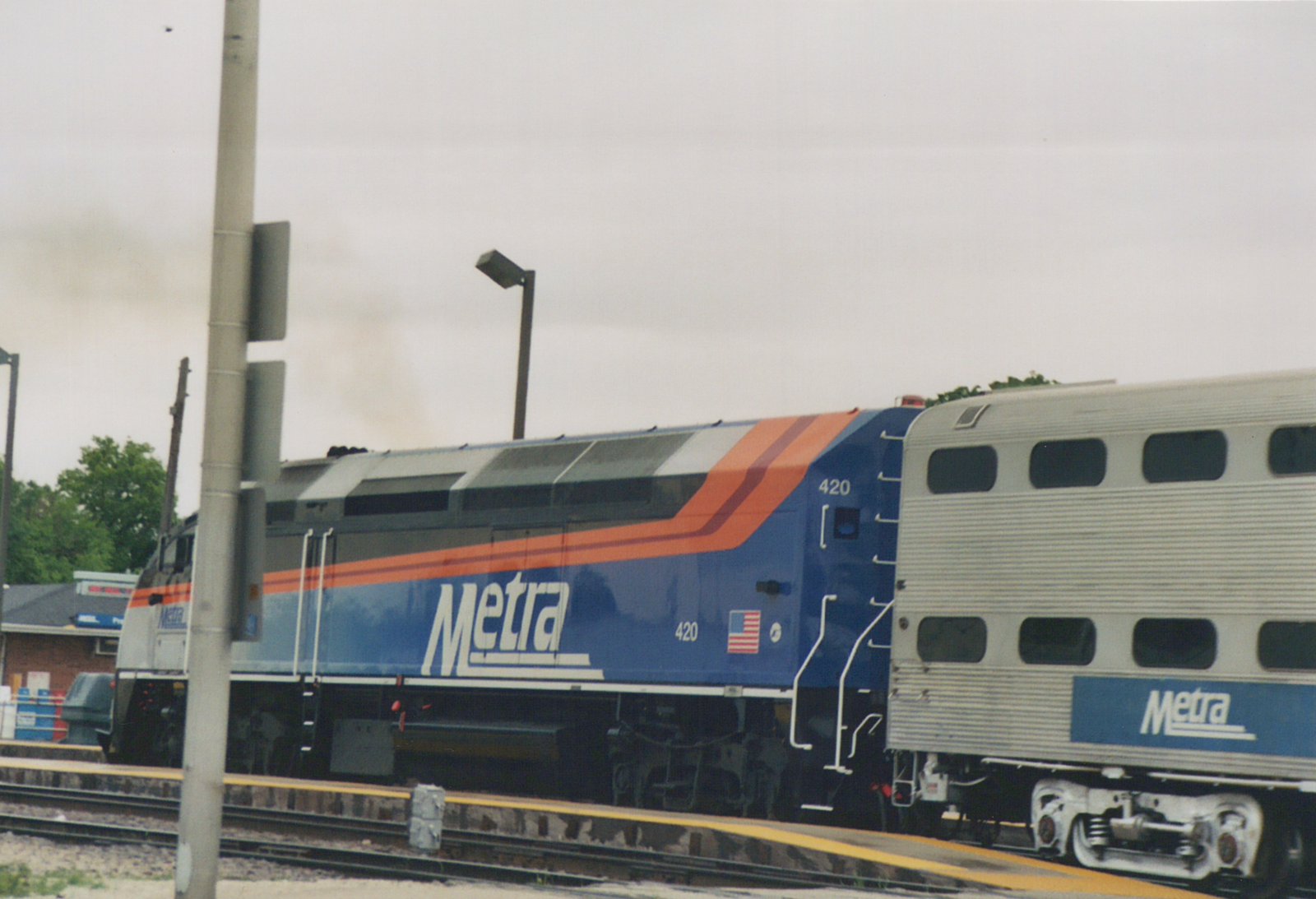A blue Metra diesel locomotive numbered 420 is parked on a track next to a station platform, pulling a silver double-decker commuter car with the Metra logo.
