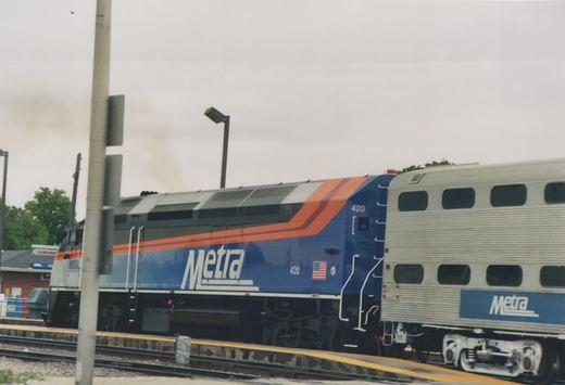 A blue Metra diesel locomotive numbered 420 is parked on a track next to a station platform, pulling a silver double-decker commuter car with the Metra logo.