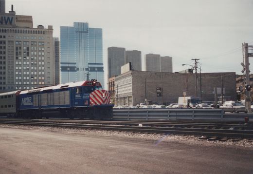 A blue and white Metra locomotive numbered 608 travels on tracks beside a street with cars and buildings in the background.