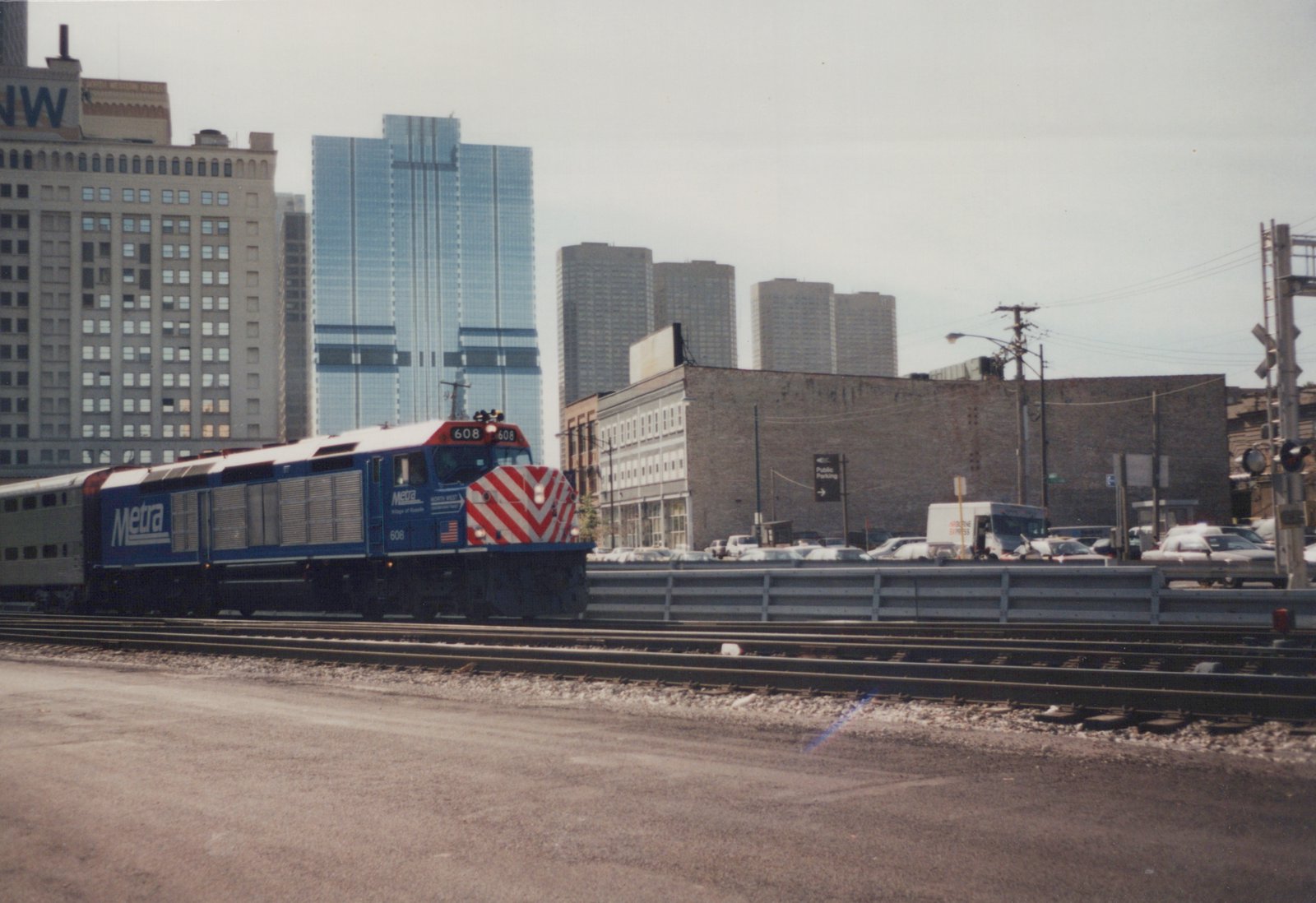 A blue and white Metra locomotive numbered 608 travels on tracks beside a street with cars and buildings in the background.