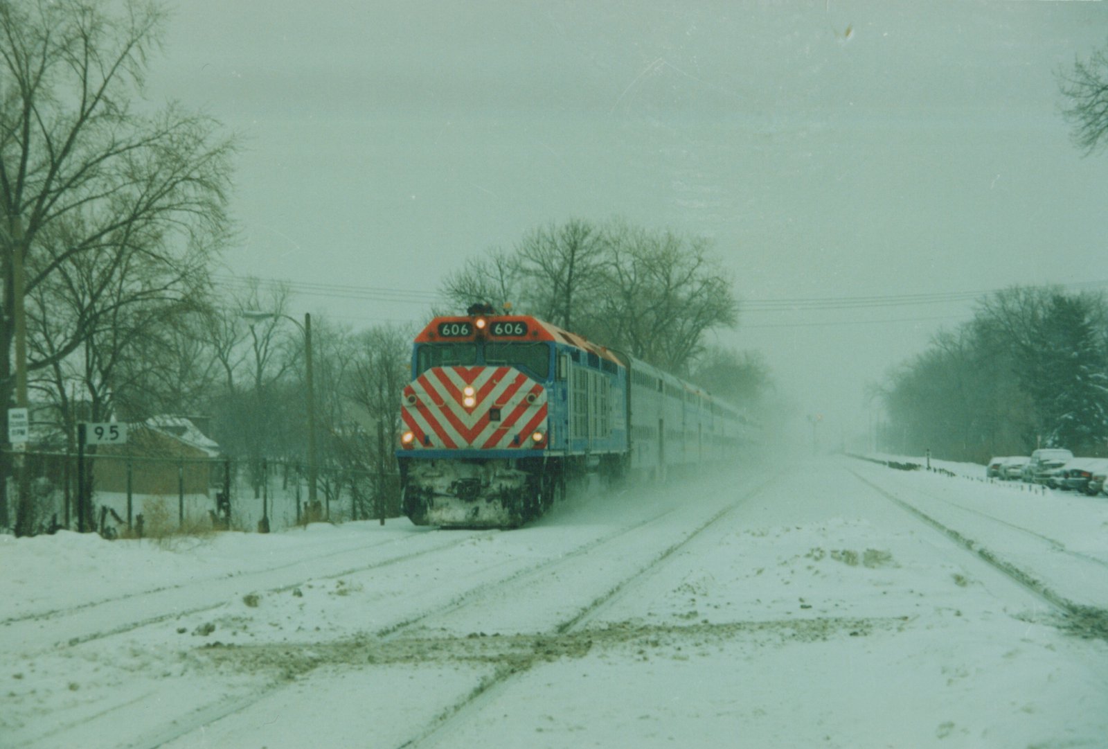 A blue and red Metra locomotive number 606 pulls a train of double-decker cars on a snowy, overcast day through a tree-lined, snow-covered rail corridor.