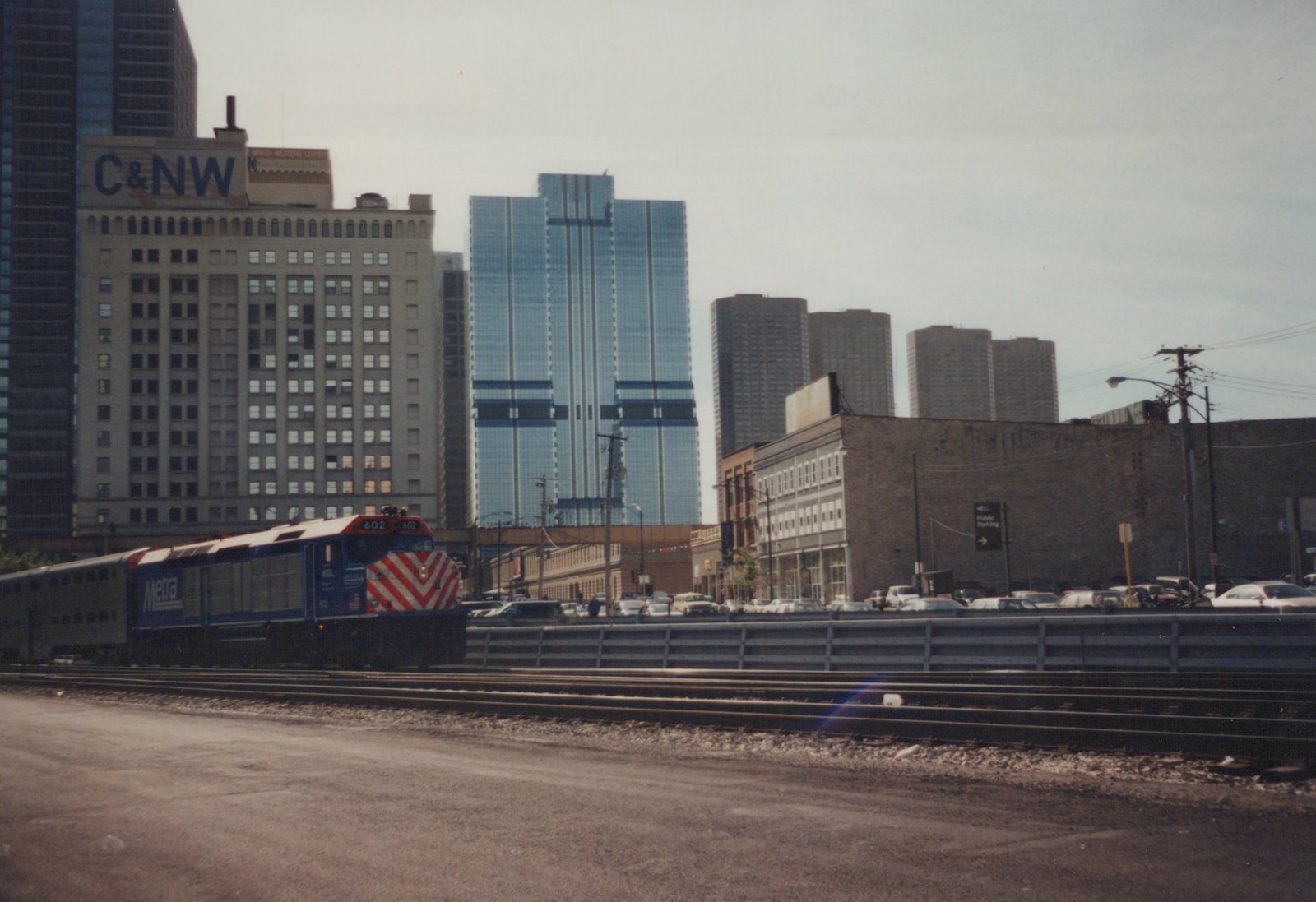 A blue Metra locomotive numbered 602 leads a train through a city scene with skyscrapers and a C&NW building sign in the background.