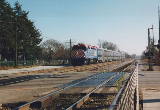 A blue and red Metra locomotive numbered 601 pulls a line of silver double-decker passenger cars across a road grade crossing under a clear sky, with trees and a fence visible in the background.