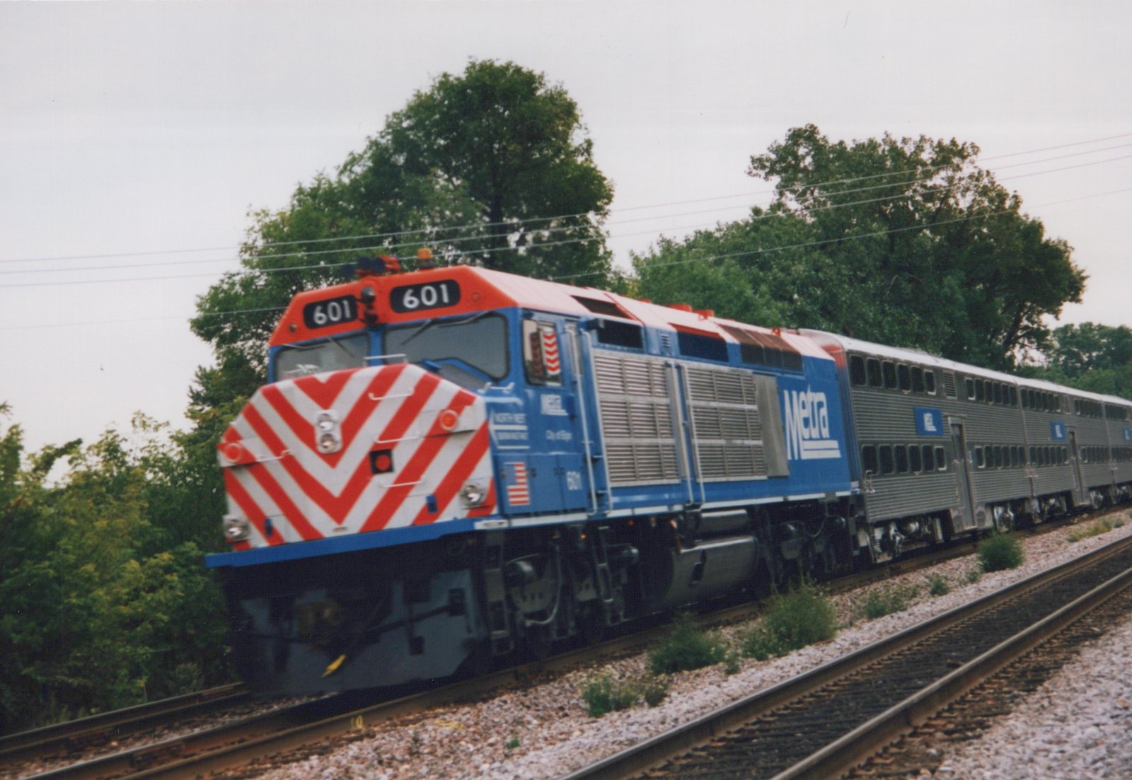 A Metra diesel locomotive pulling commuter cars on a track with trees behind under a cloudy sky.