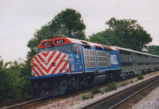 A Metra diesel locomotive pulling commuter cars on a track with trees behind under a cloudy sky.