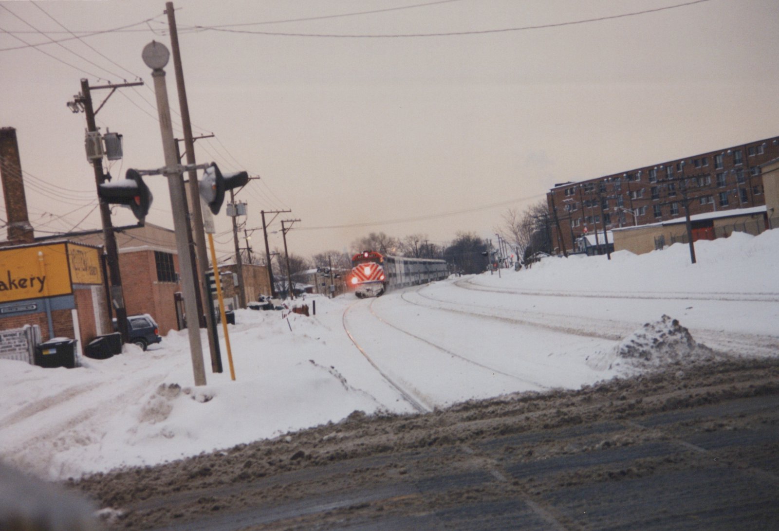 A red and white Metra locomotive numbered 607 pulls a line of silver passenger cars along snow-covered tracks beside a street and buildings.