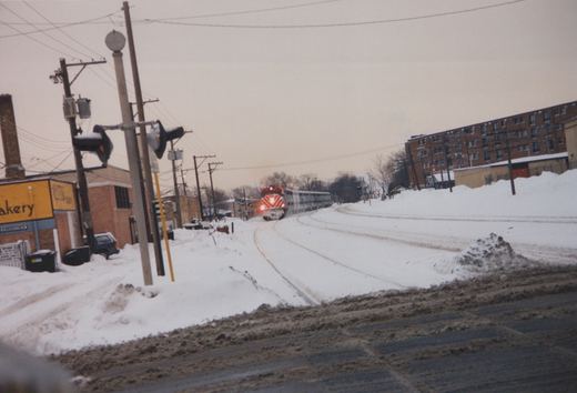 A red and white Metra locomotive numbered 607 pulls a line of silver passenger cars along snow-covered tracks beside a street and buildings.