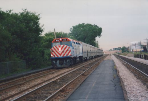 A Metra train with locomotive 611 and gallery cars travels on a private right-of-way, with trees and industrial buildings in the background on an overcast day.
