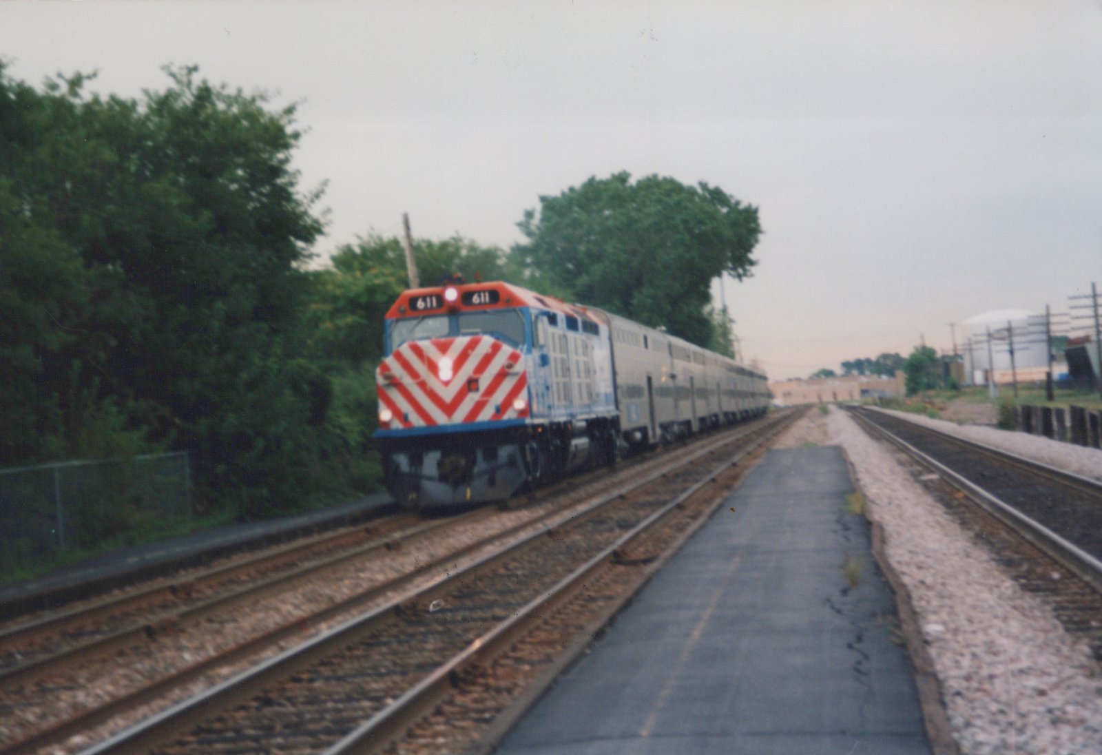 A Metra train with locomotive 611 and gallery cars travels on a private right-of-way, with trees and industrial buildings in the background on an overcast day.