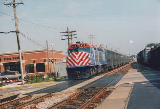 A blue and red Metra locomotive pulling silver passenger cars at a station, with a bank and a silver SUV visible in the background.