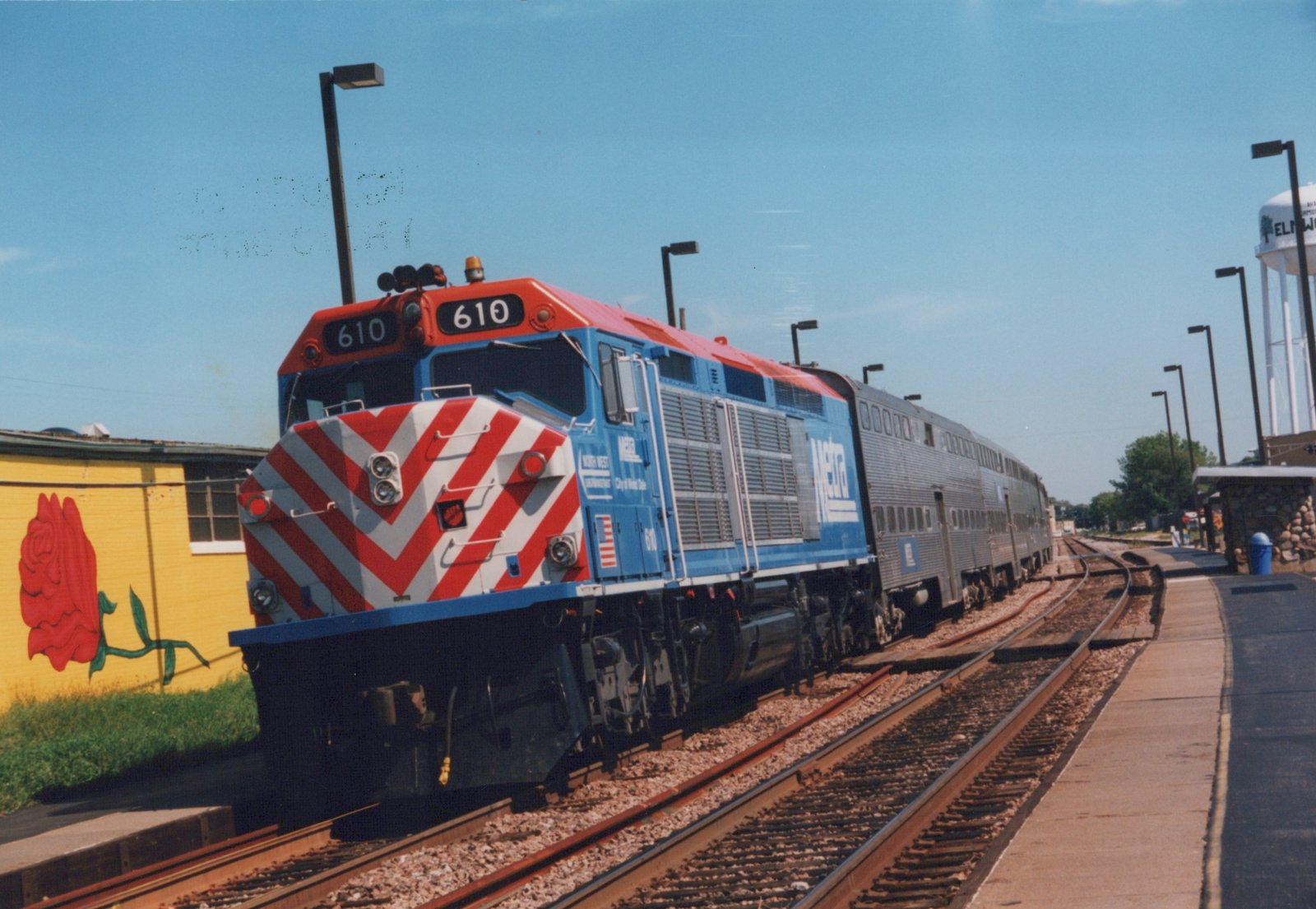 A blue and red Metra locomotive number 610 pulling silver double-decker commuter cars at a station platform, with a yellow building and a water tower in the background.