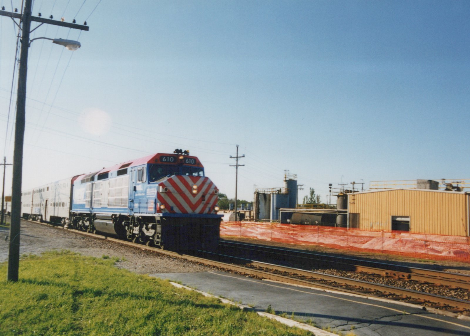 A blue and white Metra diesel locomotive with a red and white chevron nose, numbered 610, pulls a train of double-decker passenger cars along a private right-of-way next to a street with green grass and industrial buildings in the background under a clear sky.