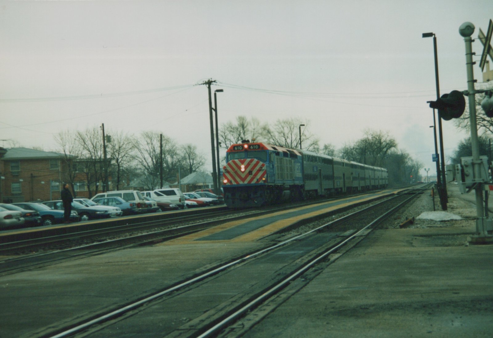 A blue and red Metra train with the number 610 on the front is stopped at a station platform with parked cars alongside and a person standing nearby.