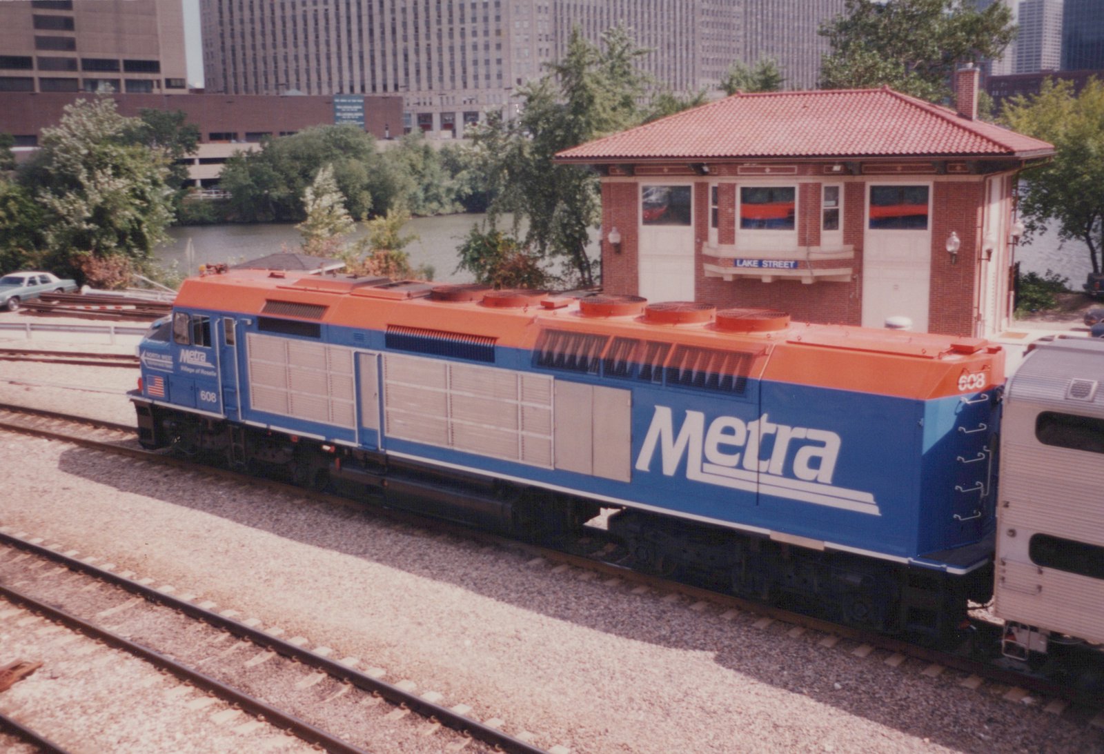 A blue and orange Metra diesel locomotive numbered 608 sits on railroad tracks next to a red brick station house labeled 'LAKE STREET', with city buildings and trees in the background.
