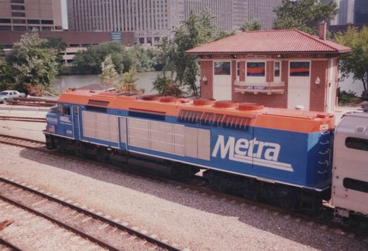 A blue and orange Metra diesel locomotive numbered 608 sits on railroad tracks next to a red brick station house labeled 'LAKE STREET', with city buildings and trees in the background.