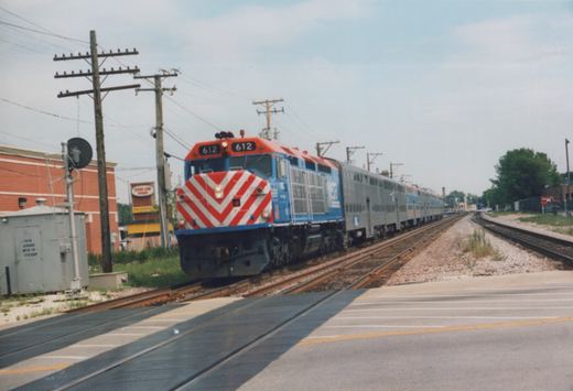 A blue and orange Metra locomotive with the number 612 on the front pulls a long train of silver double-decker passenger cars along tracks beside a road, with utility poles and a building in the background.