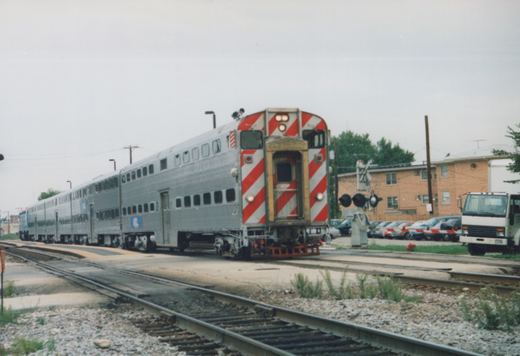 A silver Metra train of double-decker Gallery cars is stopped at a grade crossing in a residential area with a brick building and a white truck nearby.