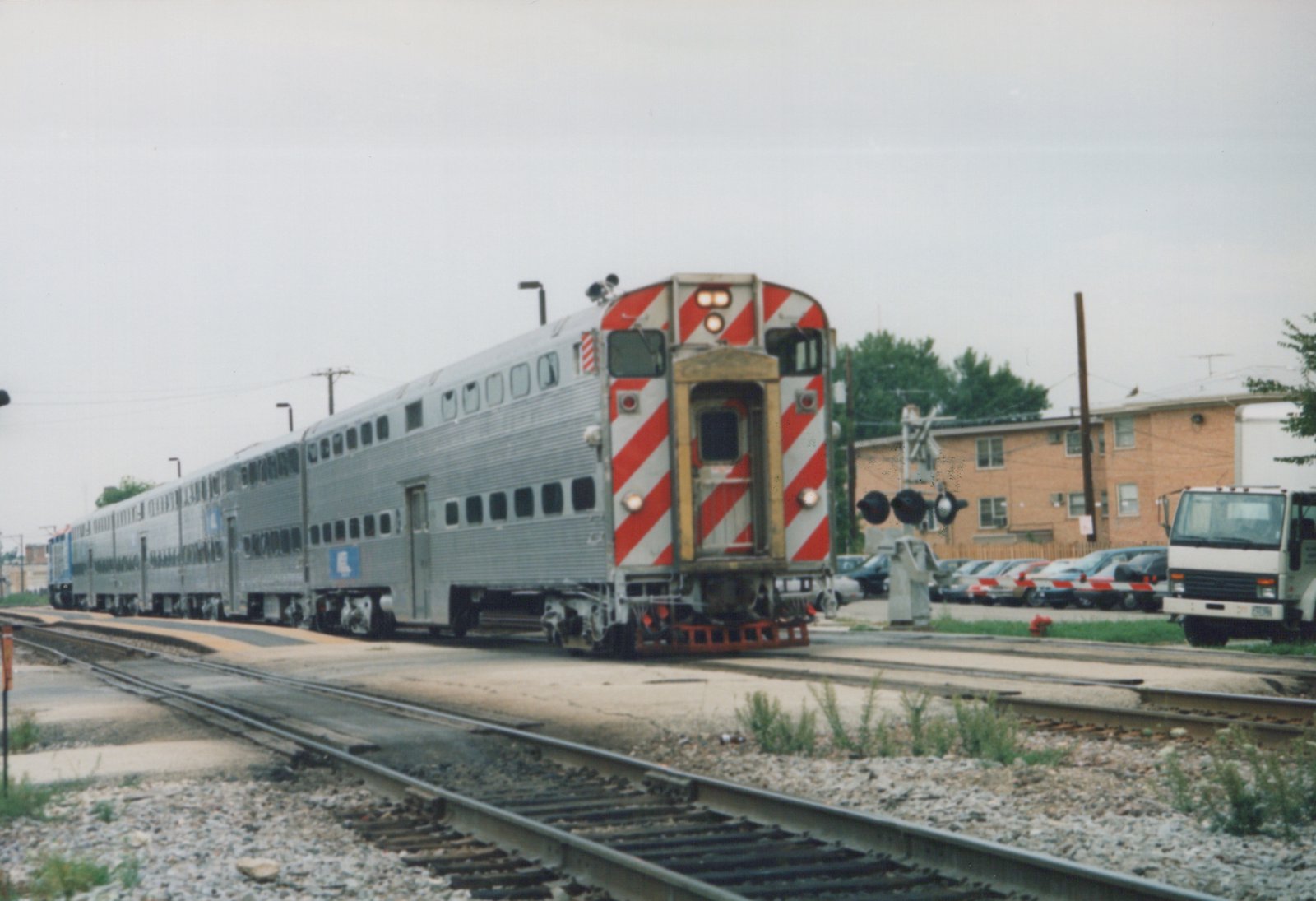 A silver Metra train of double-decker Gallery cars is stopped at a grade crossing in a residential area with a brick building and a white truck nearby.