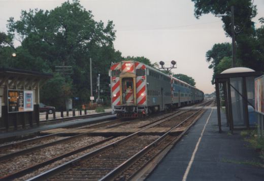 A bi-level commuter train with a silver body and red-and-white striped cab end is stopped at a station platform under an overcast sky, with trees and a shelter visible.