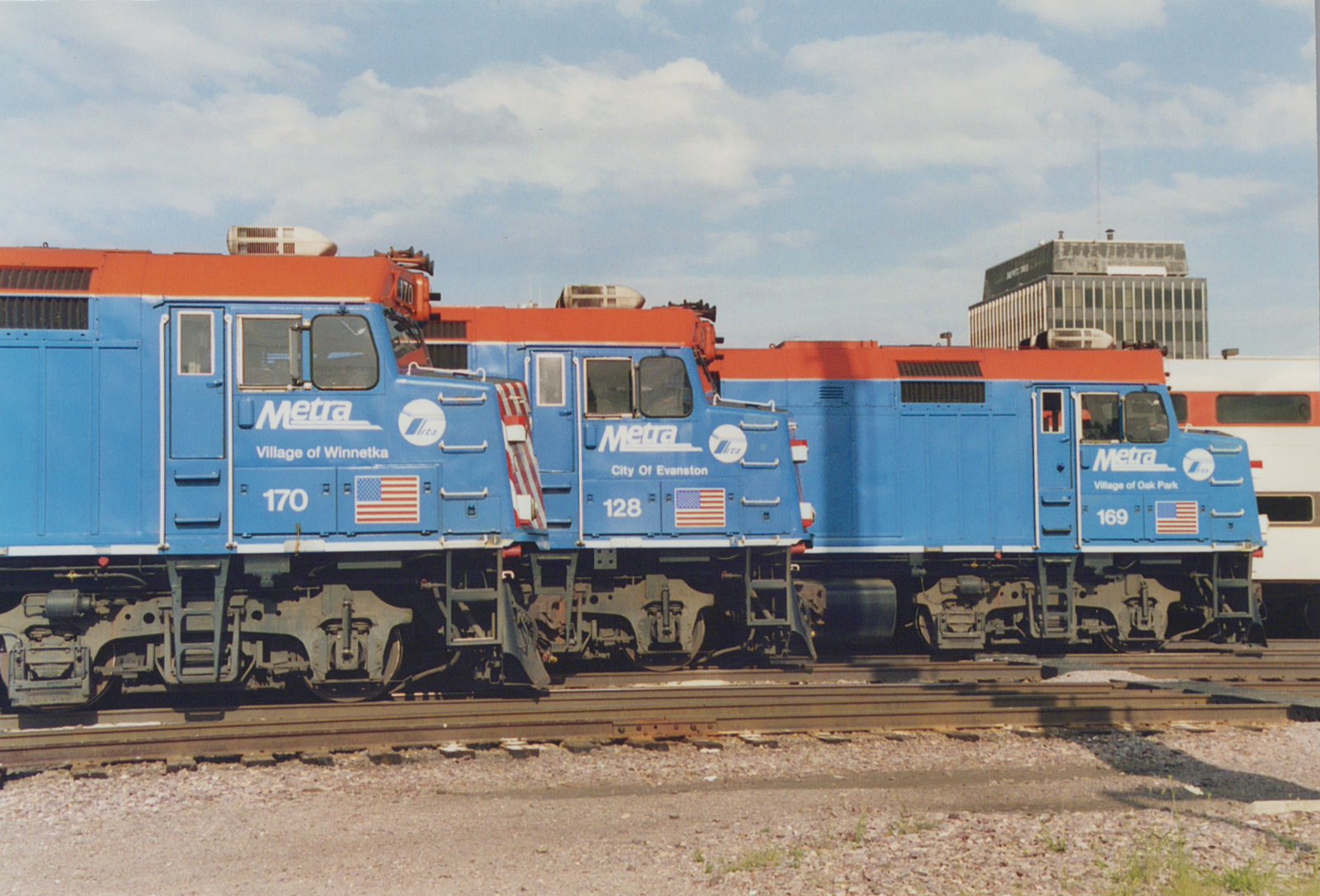 Three blue and orange Metra diesel locomotives numbered 170, 128, and 169 are parked side-by-side on tracks in a yard with a building in the background.