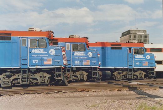 Three blue and orange Metra diesel locomotives numbered 170, 128, and 169 are parked side-by-side on tracks in a yard with a building in the background.
