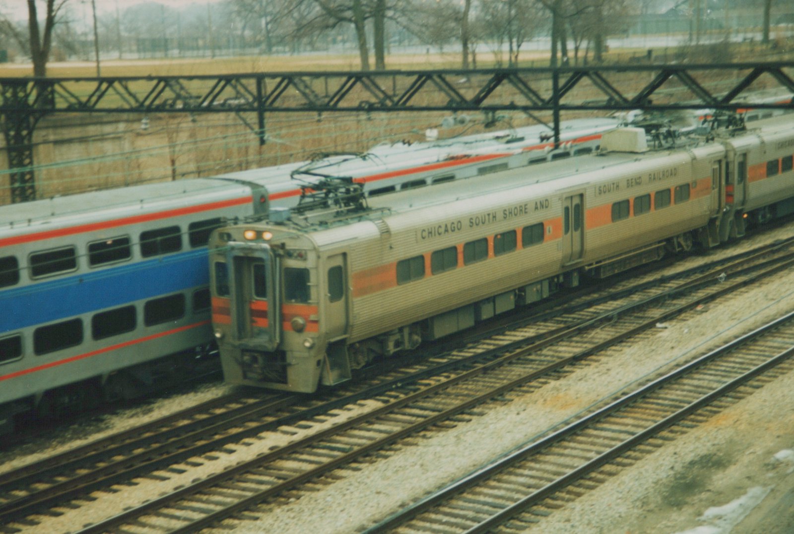 A silver and orange South Shore Line Highliner train with the number 100 on its front, traveling on parallel tracks beside a blue and red Amtrak train, under a steel truss bridge on a cloudy day with bare trees in the background.