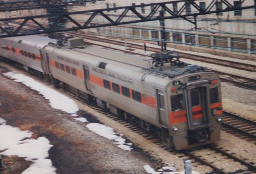 A stainless steel electric train with an orange stripe, marked 'Chicago South Shore and South Bend Railroad' and number 43, on railroad tracks near a building with overhead wires, with snow on the ground.