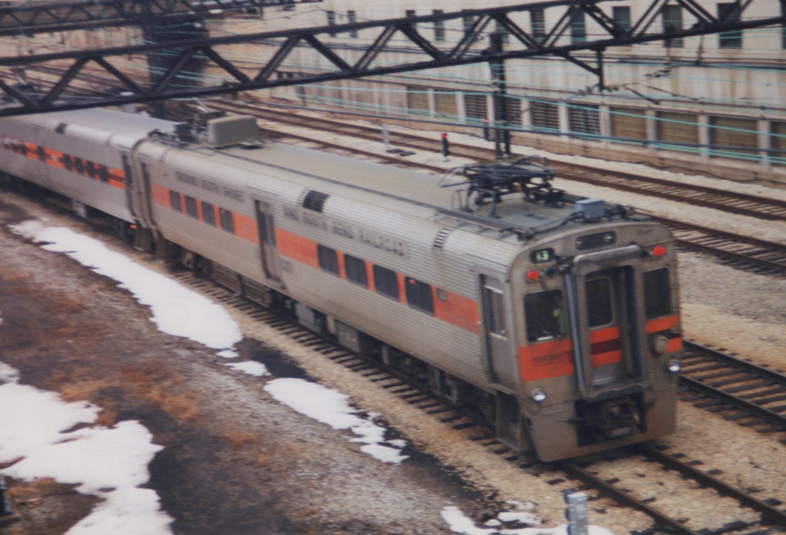 A stainless steel electric train with an orange stripe, marked 'Chicago South Shore and South Bend Railroad' and number 43, on railroad tracks near a building with overhead wires, with snow on the ground.