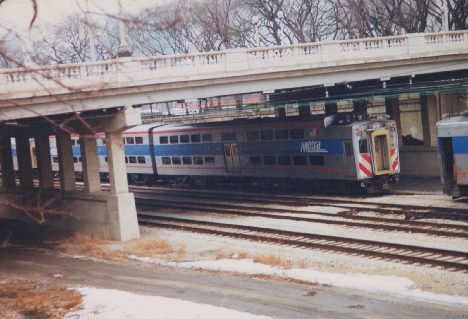 A Metra Highliner train car, numbered 758, sits on tracks under a concrete bridge with snow on the ground.